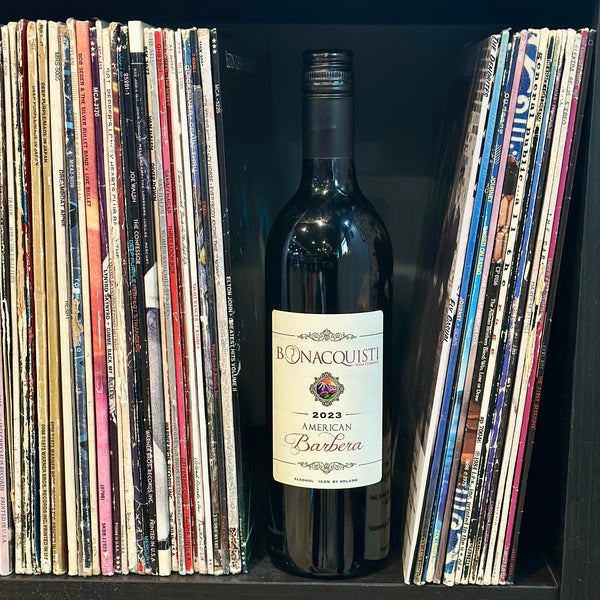 A bottle of 2023 Barbera from Bonacquisti Wine Company sitting on a bookshelf in between some vinyl records.