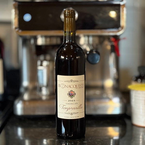 A bottle of Bonacquisti 2023 Tempranillo sitting on the counter with an esspresso machine in the background.