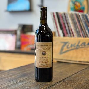A bottle of Bonacquisti 2024 Colorado Malbec red wine stands on a rustic wooden table in a record store, with vinyl records visible in the blurred background.