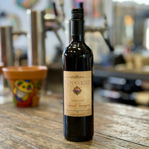 A bottle of Bonacquisti American Cabernet Sauvignon red wine stands on a rustic wooden table in a blurred bar or kitchen setting.