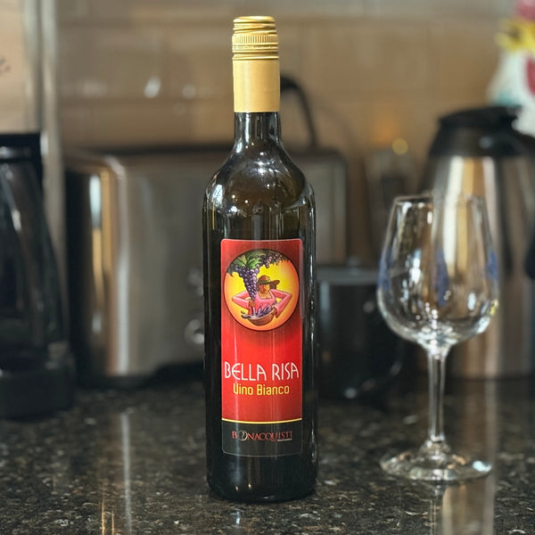 A bottle of Bonacquisti Wine Co. Bella Risa Vino Bianco on a counter with a empty wine glass. 